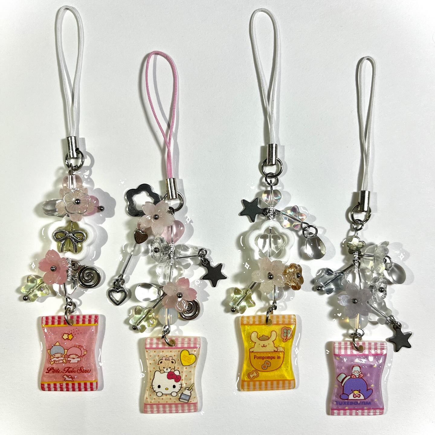 the image shows four phone charms with different cartoon characters attached to them. the charms are made of beads, flowers, and stars in various colors like pink, yellow, and purple. the cartoon characters include little twin stars, hello kitty, pompompurin, and tuxedo sam.