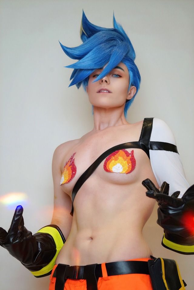 a cosplayer dressed as a firefighterthemed character with blue hair, wearing a costume consisting of black leather gloves with yellow trim, orange pants, a black strap across the chest, and fireshaped appliques covering the nipples. she is posed with her hands up and displaying her midriff and abs.