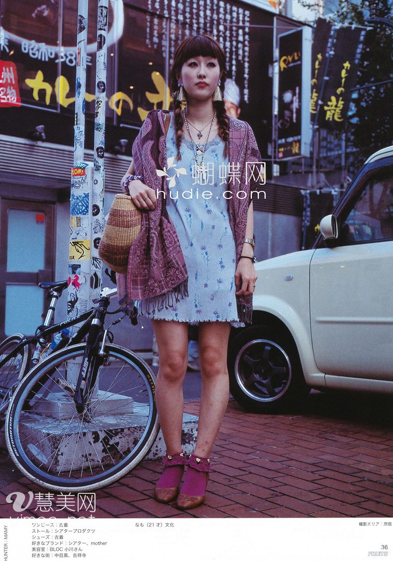 a young woman is standing on a city street, posing for a street style photo. she's wearing a light blue floral dress, a purple shawl, and purple ankle boots. she's also carrying a woven bag. a bicycle is leaning against the wall behind her. the image has a slightly faded, vintage look.