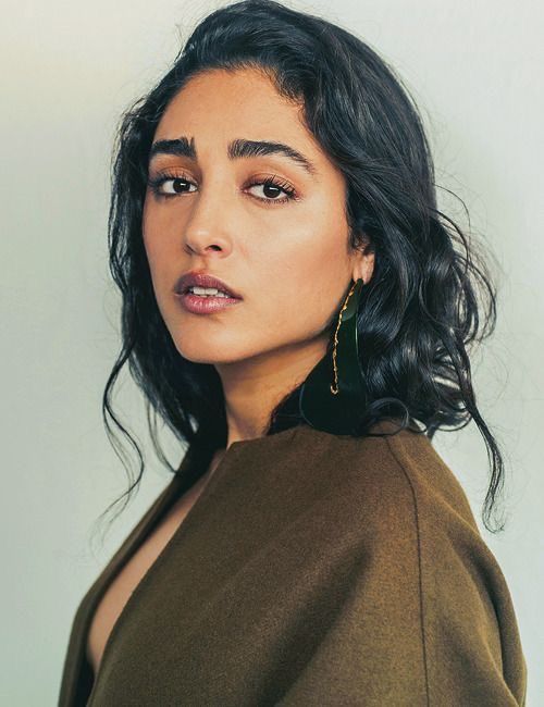 a woman with dark hair and dark eyes is wearing a brown coat and a large, darkcolored earring. she is looking at the camera with a slightly serious expression.