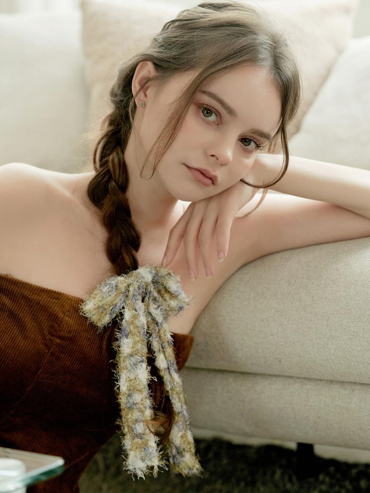 a woman with brown hair in a braid with a fabric bow is leaning against a couch. she is wearing a brown corduroy dress.