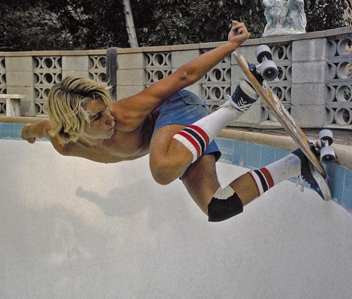 a young man in a blue denim cutoff shorts and striped socks does a trick while skateboarding in a pool. he is in mid air, with his board perpendicular to the pool wall and his body is bent in a curve, his right hand is up high by his head and his left hand is holding the board. the pool has tile trim at the edge, and a cement wall behind. there are trees and a fence in the background.