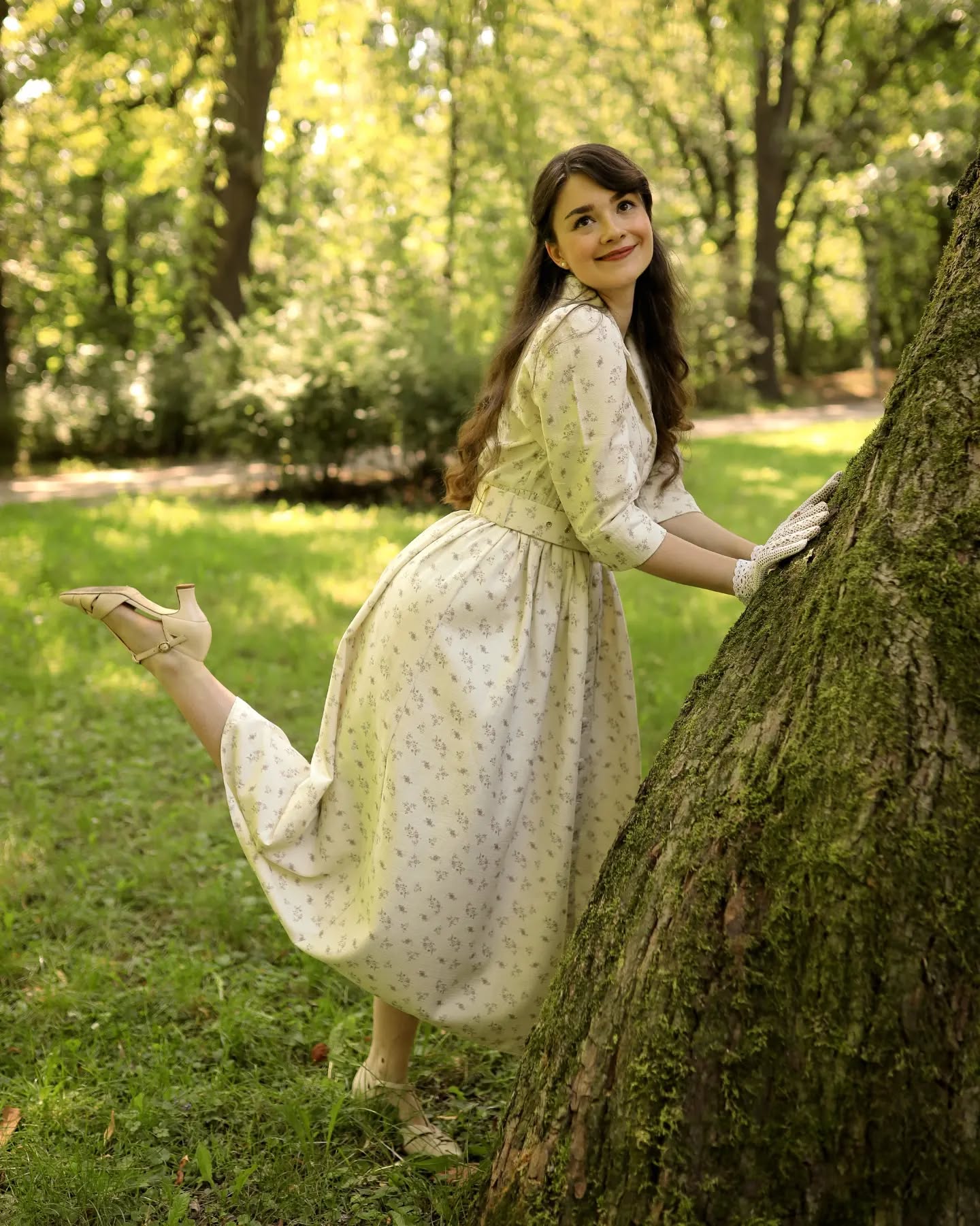 a young woman in a light beige floral vintage dress is playfully leaning against a large tree in a park. she is smiling and wearing light beige heels and white gloves. the sunlight filters through the trees creating a dreamy ambiance.  the scene is idyllic and suggests a sense of carefree joy.