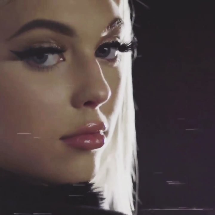 a closeup portrait of a woman with striking makeup. she has defined eyeliner, long eyelashes, and glossy lips. her blonde hair is visible on the side. the background is black, emphasizing her face and features.