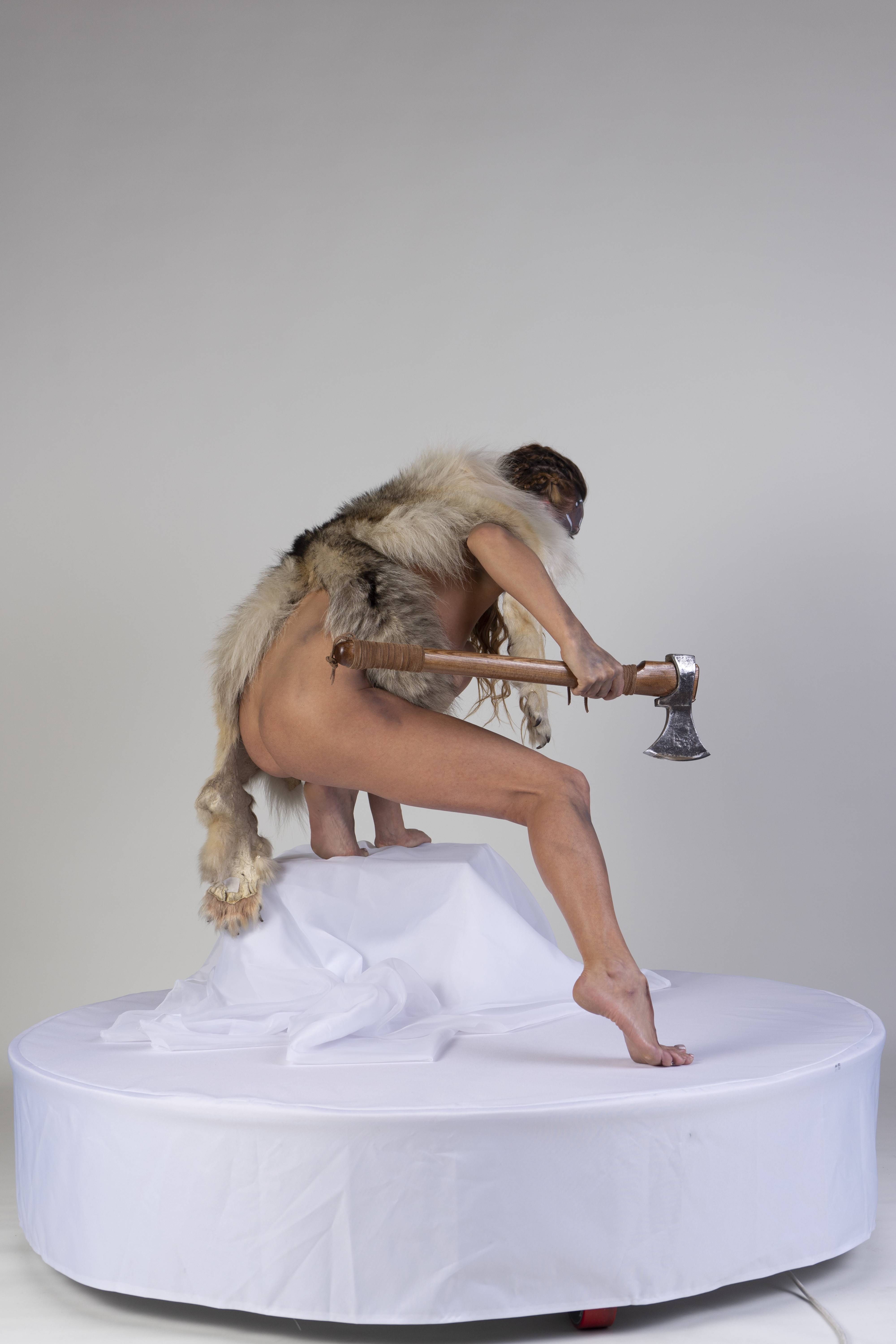a nude woman wearing a fur pelt and holding an axe stands on a white platform against a white background.