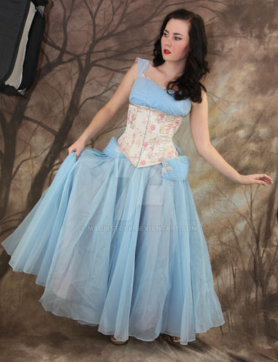 a fulllength studio shot of a young woman wearing a beautiful light blue tulle gown over a pale pink and white floral corset. the dress has a full, flowing skirt and a simple, elegant bodice. the woman is posing gracefully against a backdrop of stylized trees, creating a romantic and dreamy atmosphere reminiscent of a fairytale or vintage film. the overall aesthetic is vintageinspired, possibly evoking the 1940s or 1950s. her dark hair is styled in a slightly wavy manner, and her makeup is subtle but sophisticated. the image is welllit and the colors are vibrant and harmonious. the watermark "magikstone" is visible on the dress.