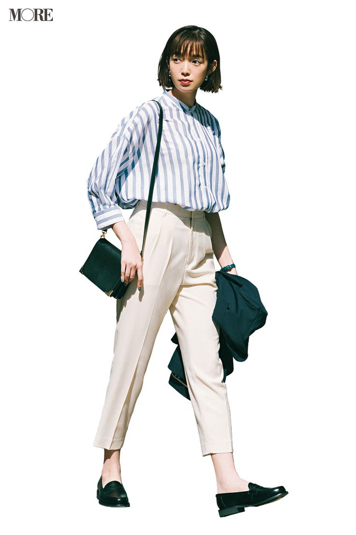 a young woman in a striped shirt, white pants, and black loafers is walking down the street. she is carrying a black bag on her shoulder and has a jacket draped over her arm. her style is trendy and casual. she is looking to her left, and her expression is relaxed.