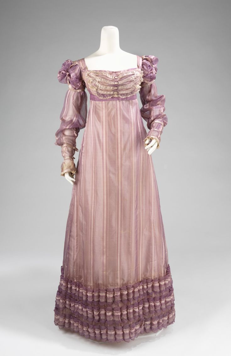 a fulllength, longsleeved pink and purple striped dress with lace ruffles around the neckline and waist. the dress is gathered at the waist and falls to the floor in a long, flowing skirt. the fabric is sheer and translucent, and the dress is trimmed with lace and ruffles. the dress is a beautiful example of early 19thcentury fashion.