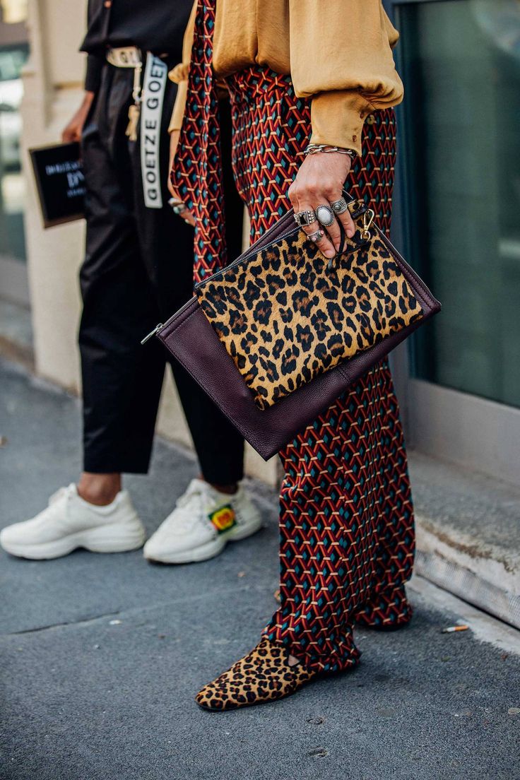 the image shows a woman's street style outfit. she's wearing red and blue geometric pattern pants, leopard print shoes, and a brown shirt. she's also carrying a leopard print clutch and wearing rings on her fingers. the style is autumnal and trendy. another person wearing sneakers and black pants is visible in the background. the setting appears to be an outdoor street.