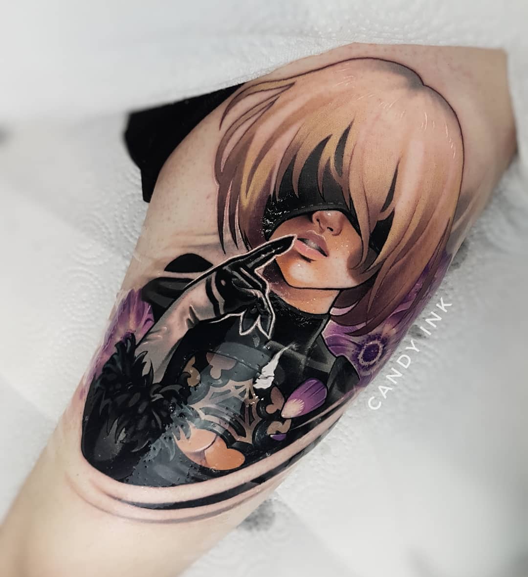 a closeup photograph showcasing a vibrant tattoo of the character 2b from the video game nier automata. the tattoo, executed in a neotraditional style, is inked on the upper arm of an individual. the character is portrayed in her signature black outfit, adorned with floral accents, and her iconic blindfold. the artwork demonstrates a blend of blackwork and color, resulting in a visually striking piece.