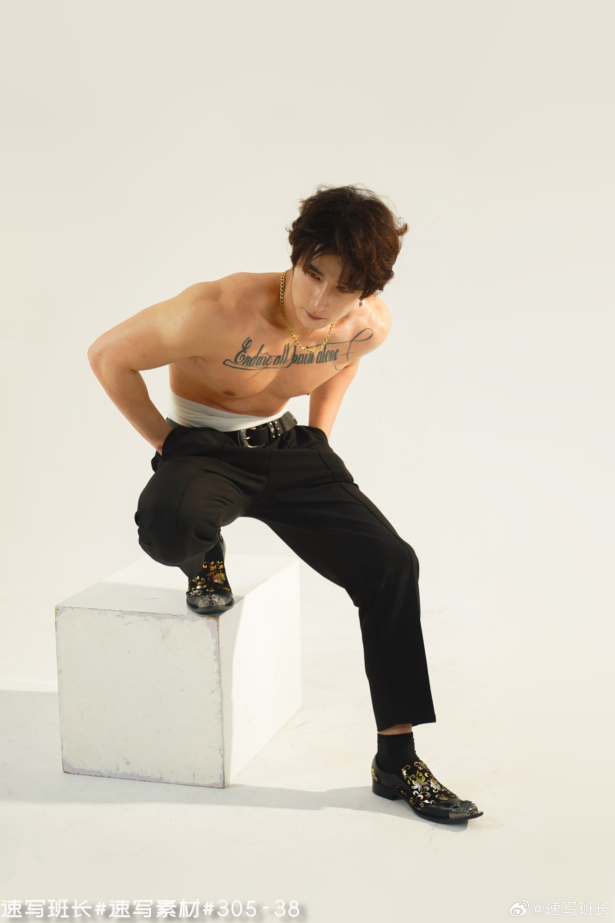 a shirtless asian man with a tattoo on his chest is posing in a studio. he's wearing black pants and stylish shoes with a gold pattern. he's leaning on a white cube with his hands in his pockets, looking down. the background is entirely white and brightly lit.