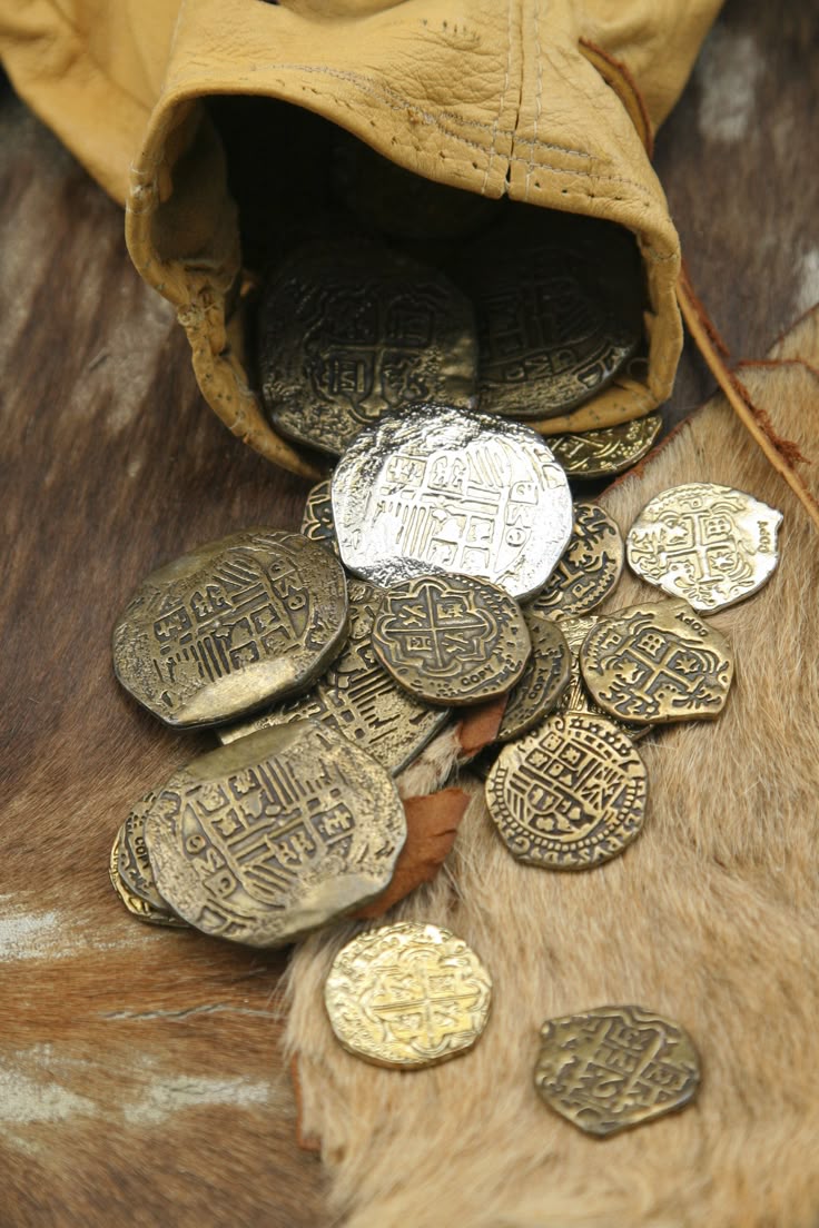 an image of a leather bag spilling out various antiquelooking coins onto a furry surface. most of the coins are a gold color, with one prominent silver coin among them. the bag itself is made of a tancolored leather.