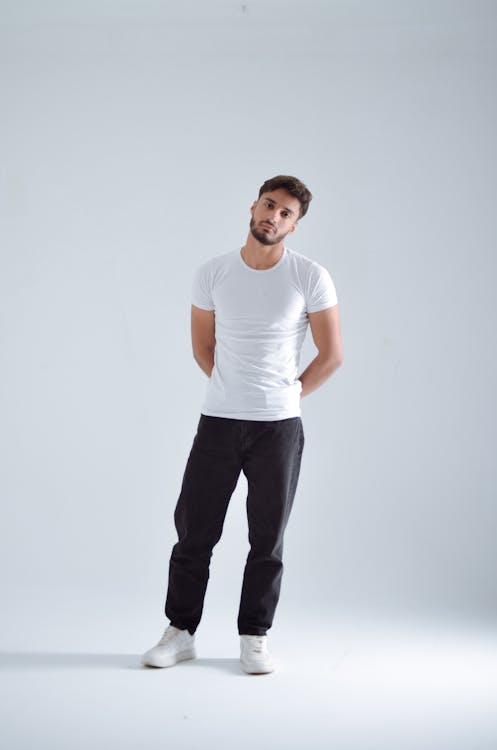 a full body shot of a man in a studio setting. he's wearing a white tshirt, black jeans, and white sneakers. he has a beard and is standing in a casual pose with his hands behind his back. the background is a light grey/white.
