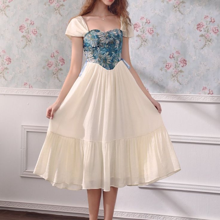 a woman is wearing a vintagestyle, romantic dress. the dress is ivory with a floral bodice and a flowing skirt. the dress has short puff sleeves and a square neckline. the woman is standing in front of a floral wallpapered wall. the lighting is soft and the perspective is at eye level.
