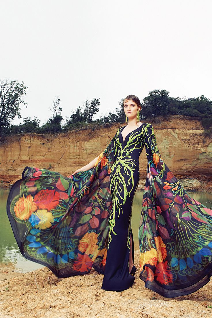 a woman wearing a long, floral print dress with a flowing train and wide sleeves, is standing in a natural setting, with rocks, water, and greenery behind her. she is looking away from the camera.  she is posing in a glamorous fashion style. the dress is detailed with an intricate  design that is reminiscent of nature, with green vines and flowers. the model's confidence and the beautiful landscape add to the overall impact of the image.