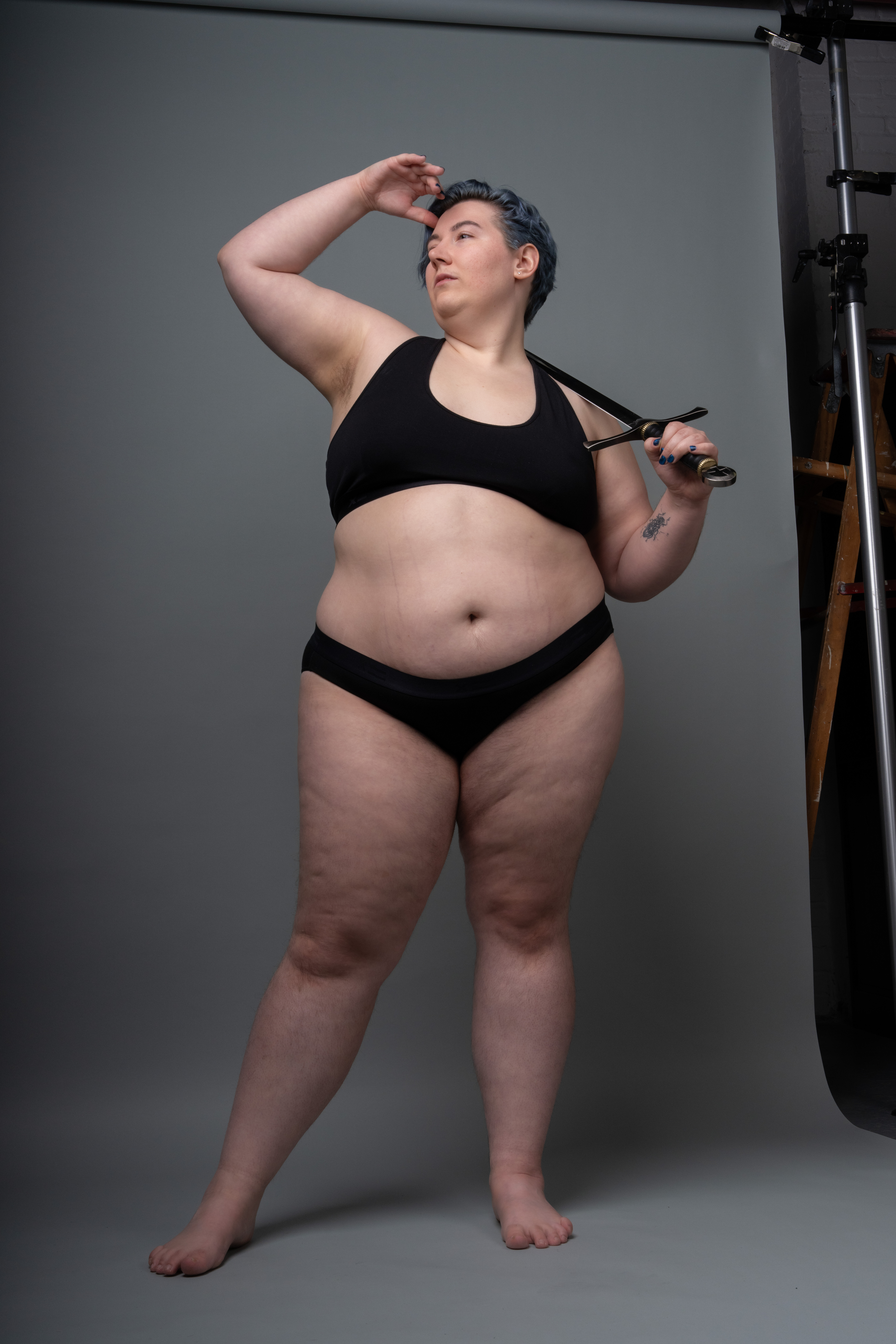 a plussize woman with blue hair is posing in a studio. she is wearing a black sports bra and black underwear. she is holding a sword over her shoulder with her right hand and has her left hand on her forehead, as if shielding her eyes. she is standing barefoot on a grey background. there is a light stand partially visible on the right side of the frame.