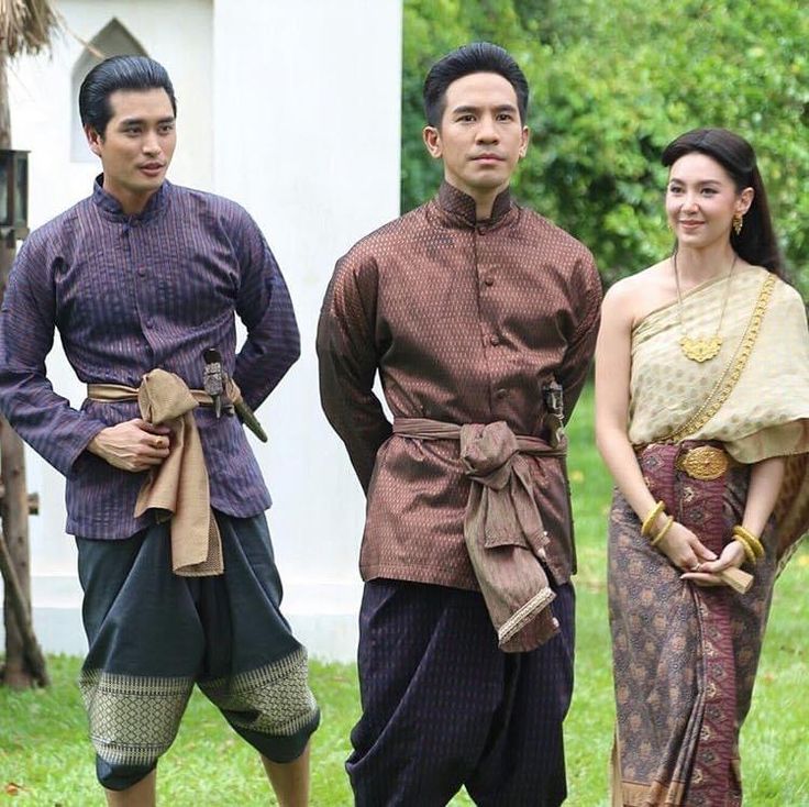 three people, two men and one woman, are wearing traditional thai clothing. the two men are standing side by side, while the woman is standing to their right. the men are both wearing longsleeved shirts and long pants, while the woman is wearing a sleeveless top and a long skirt. all three of them are smiling. the men's shirts are decorated with intricate patterns, and their pants are made of a fine, silky material. the woman's top is made of a sheer fabric, and her skirt is adorned with colorful embroidery. the background of the image is a lush, green garden.