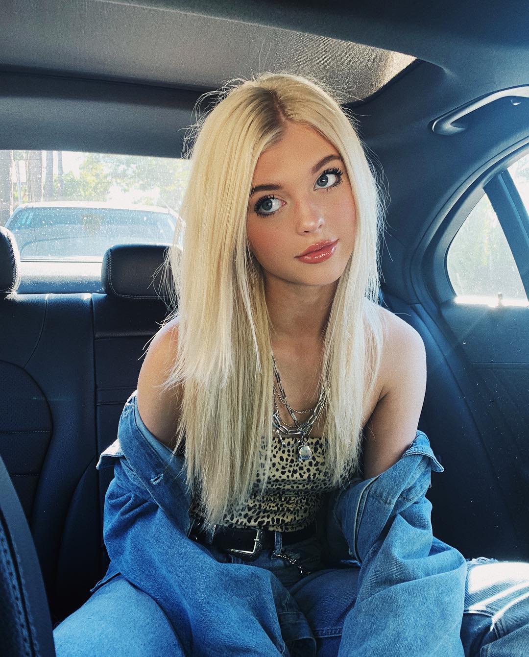 a young blonde woman with blue eyes and lip gloss is sitting in the backseat of a car. she is wearing a leopard print top, a denim jacket, and a layered necklace. the car's interior is visible, with the backseat and headrest in focus.