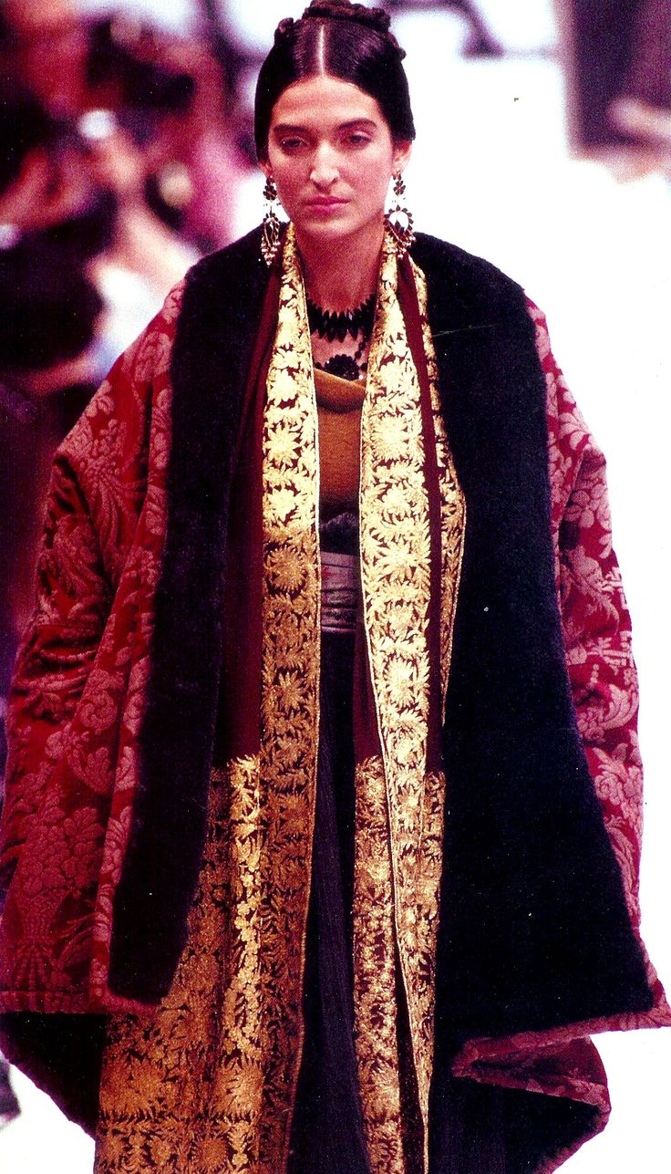 a woman model is shown on a runway. she is wearing a long, richly embroidered coat with a fur collar. the coat is dark red velvet with gold embroidery. the model is wearing a gold necklace and earrings. the background is blurred but shows an audience.  the image is vintage, possibly from the 1990s. the overall style is elegant and sophisticated.
