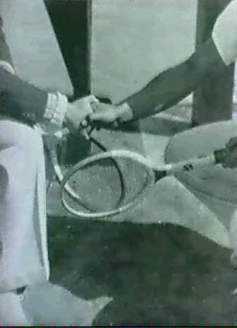 black and white vintage photograph shows two tennis players shaking hands on a tennis court. one player is seated, while the other is standing.  the players are close together, with their hands clasped. a tennis racket rests between them. the image has a grainy texture and a somewhat muted color palette. the focus is slightly soft, but the overall scene is clear enough to identify the subject matter.
