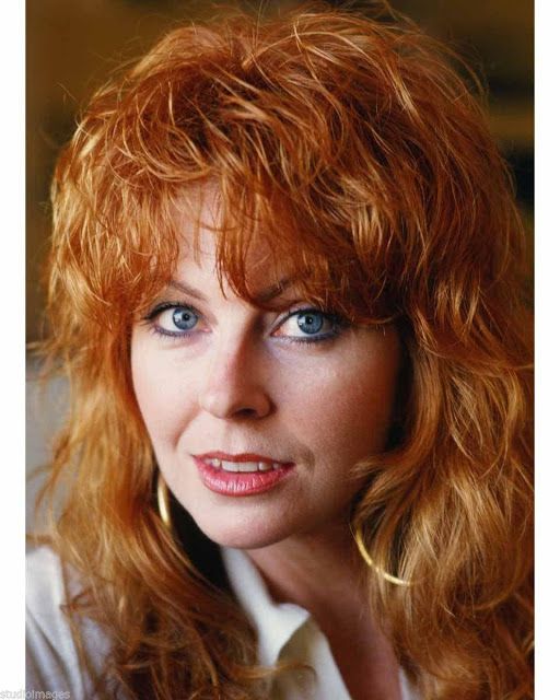 a closeup headshot of a redheaded woman with long, wavy hair, smiling slightly at the camera. she's dressed in a simple, casual white shirt. the background is a blurry, out of focus.  she looks like she's from the 70s or 80s.