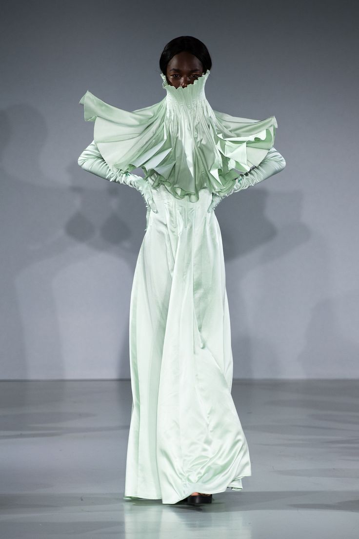 a slender, darkskinned model stands on a pale gray runway, showcasing a dramatic, light green couture gown. the gown features a high, pleated neck that conceals the lower face. flaring out from the neck are ruffled, winglike structures extending to the shoulders. the dress has long sleeves with matching gloves, and the lower portion flows into a wide, elongated skirt that trails slightly on the floor. the model's hands are placed on her waist, accentuating the dress's form. the background is a minimalist, light gray, providing a clean and focused environment for the fashion presentation.