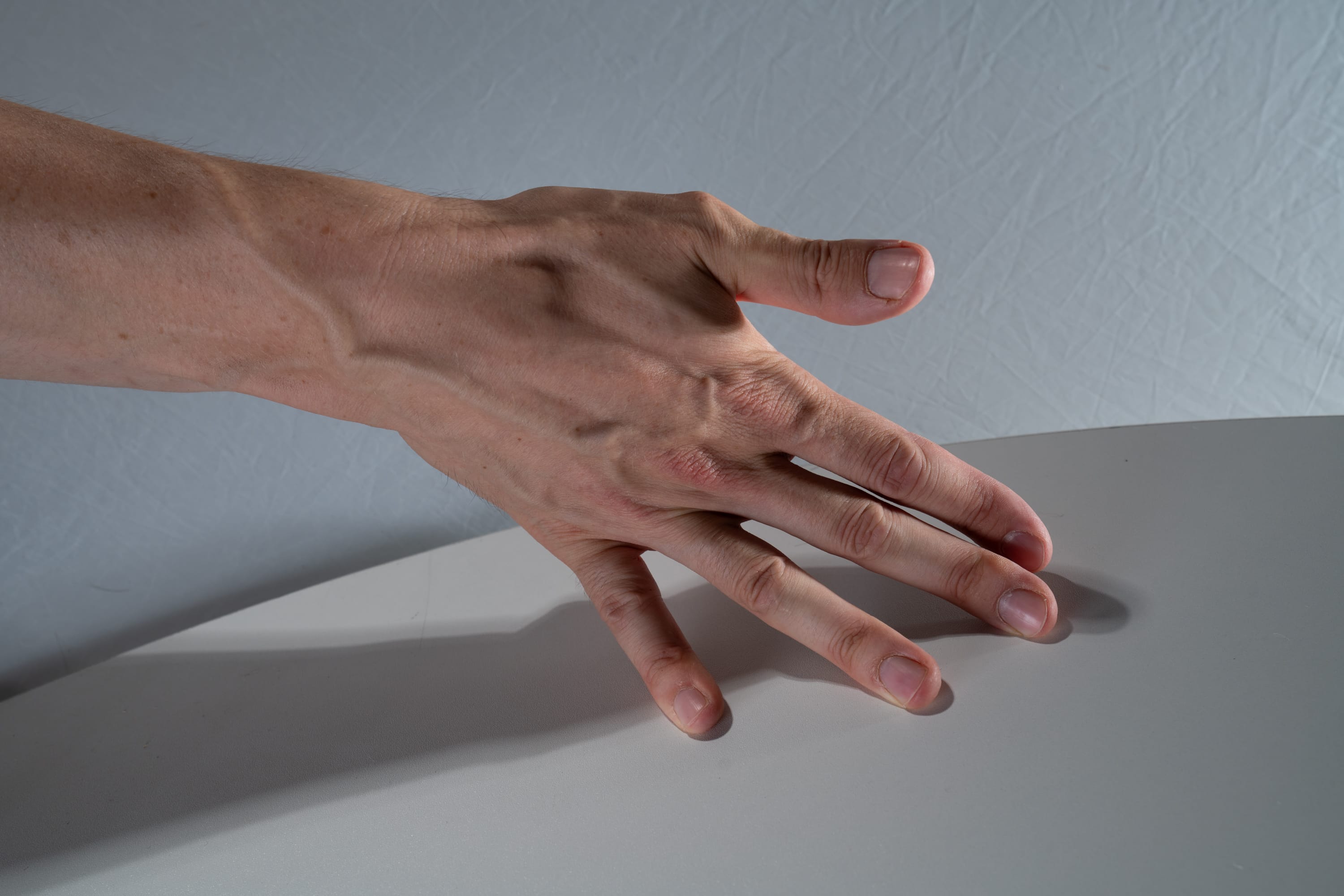 a person's hand is extended, palm down, with fingers spread slightly, resting on a white table.  the hand is positioned with the pinky side closest to the camera and the thumb furthest away. the light source is from the upper left, creating shadows from the hand onto the table.  veins are prominent on the back of the hand.