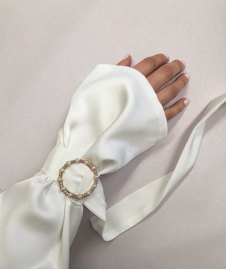 a closeup shot of a hand and wrist adorned with a white fabric sleeve featuring a pearlembellished accessory. the sleeve is gathered or tied around the wrist with a white ribbon and fastened with a circular buckle adorned with pearls. the hand is delicate with neatly manicured nails, and the fabric is smooth and luxurious, catching the light softly.