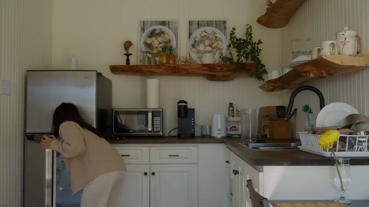 a woman is reaching into a stainless steel refrigerator in a bright, naturally lit kitchen.  the kitchen features white cabinets, dark countertops, and open wooden shelves decorated with plants and kitchenware. a microwave, coffee maker, and other kitchen essentials are neatly arranged on the counter.