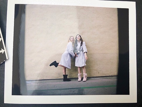 the image is a polaroidstyle photograph depicting two teenage girls standing against a beige wall outdoors.  both girls are wearing light purple or lavender bathrobes and uggstyle boots. one girl wears darkcolored boots, while the other wears light pink ones. they appear to be posing casually, one slightly leaning against the other. the photo has a slightly grainy quality characteristic of film photography, adding to its vintage aesthetic. the background is a simple, plain wall, drawing focus to the two subjects. the overall mood is relaxed and friendly.