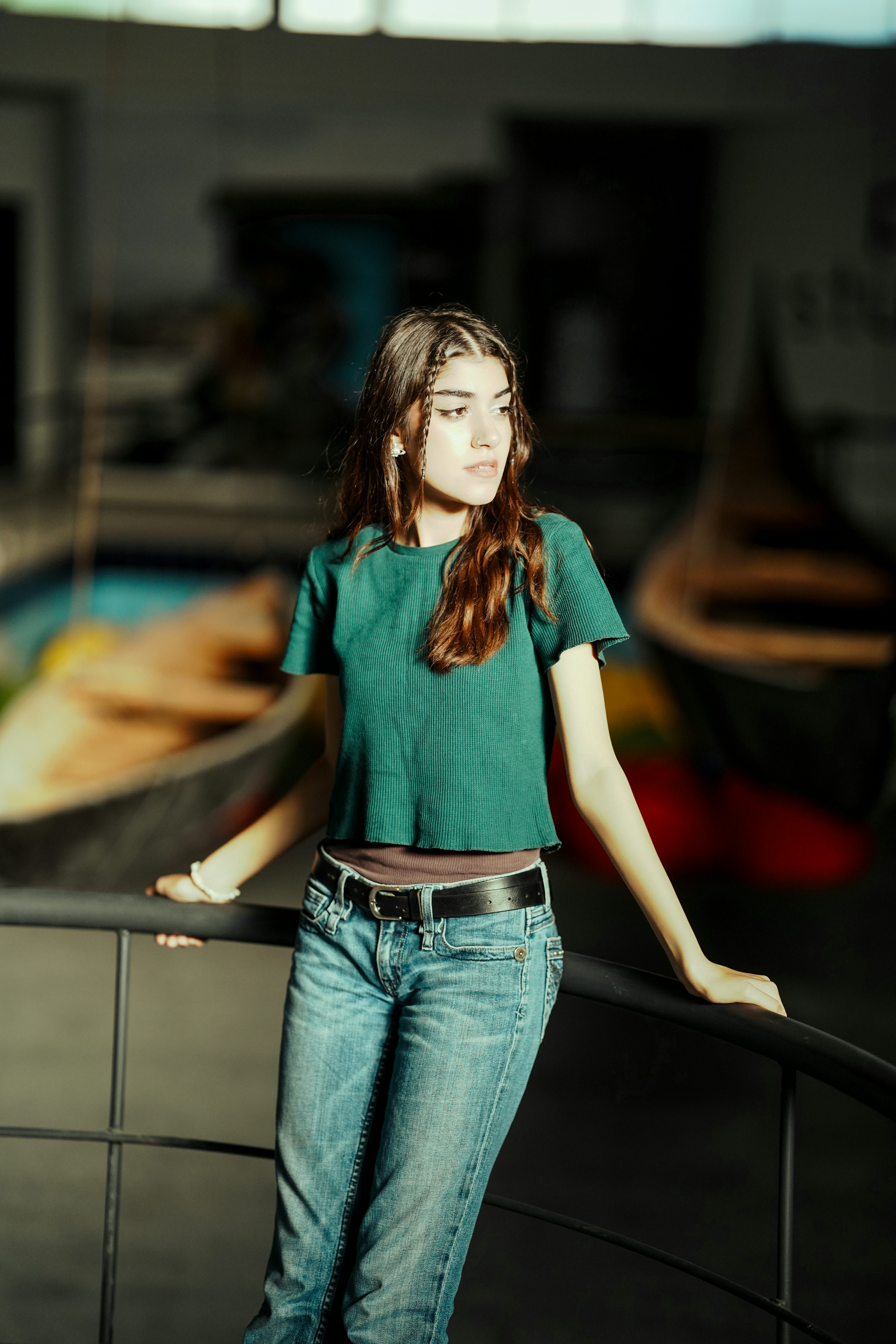a young woman in jeans and a tshirt leans against a black metal rail while posing for a photo. she has long brown hair with braids near her face. she wears a black belt, a brown undershirt, and a white wristband. the background features blurred images of boats and an indoor setting.