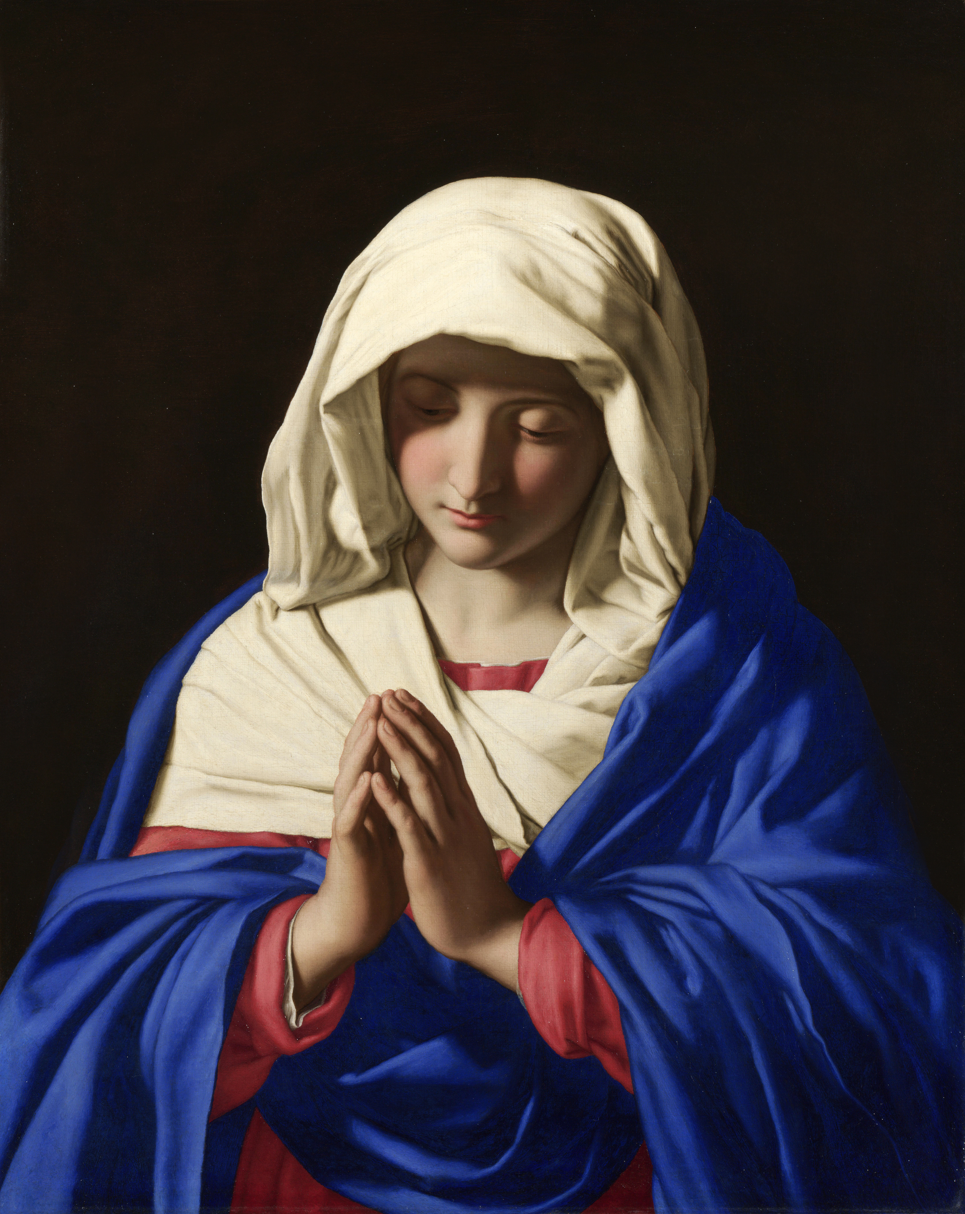 the image is a painting of the virgin mary. she is depicted in prayer, her hands clasped together, and her head bowed in contemplation. she is wearing a blue robe over a red garment and a white veil. the background is dark, emphasizing the figure of the virgin mary. the painting is likely a depiction of the virgin mary's piety and devotion to god. the image evokes a sense of peace, serenity, and faith.