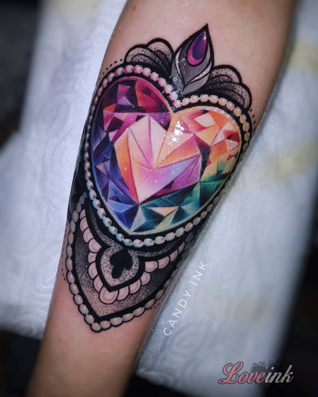 a tattoo of a colorful, jewelencrusted heart on someone's forearm. the tattoo is highly detailed and realistic, with intricate patterns and shading. the heart is the focal point of the tattoo and is surrounded by a decorative border. the tattoo is on the left forearm and is partially covered by the person's hand. the tattoo artist's signature, "candy ink," is visible in small letters below the heart.