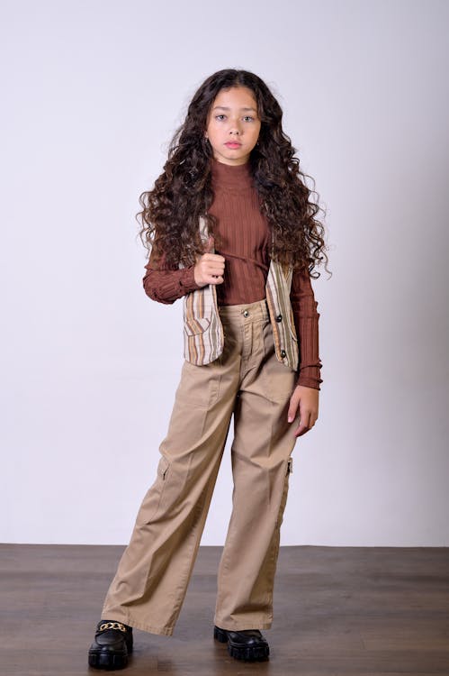 a young girl with curly hair is posing in a studio. she is wearing a brown turtleneck shirt, a striped vest, and tan cargo pants. she is also wearing black shoes. she is looking directly at the camera.