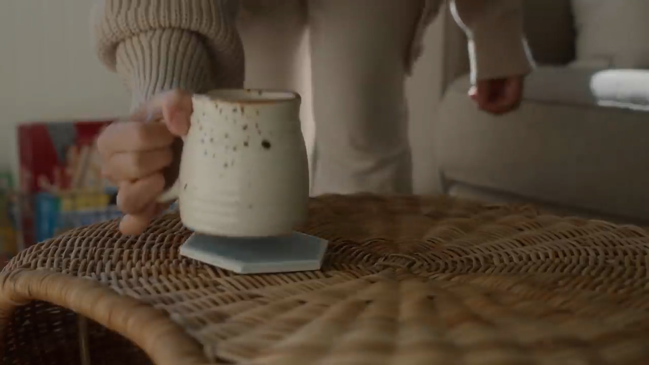 a woman is placing a mug of coffee on a woven table. the mug is white with brown speckles and is resting on a light blue hexagonal coaster. the table is made of natural wicker, and the woman is wearing a cozy beige sweater. the lighting is soft and natural, creating a warm and inviting atmosphere.