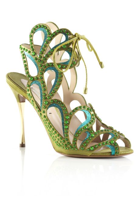 a closeup shot of a pair of elegant women's highheeled sandals. the shoes are a light olive green satin with intricate cutouts and embellished with numerous small green and teal crystals. the thin gold heels are also visible. the shoes are presented against a white background, and their reflection is slightly visible beneath them.  the sandals have a laceup detail around the ankle.