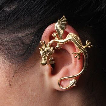 the image shows a closeup of an ear wearing a gold dragonshaped ear cuff. the ear cuff wraps around the ear, with the dragon's head facing forward and its body and tail curving around the ear. the dragon has detailed scales, wings, and claws. the background shows dark hair and skin. the jewelry is ornate and appears to be made of a shiny metal.