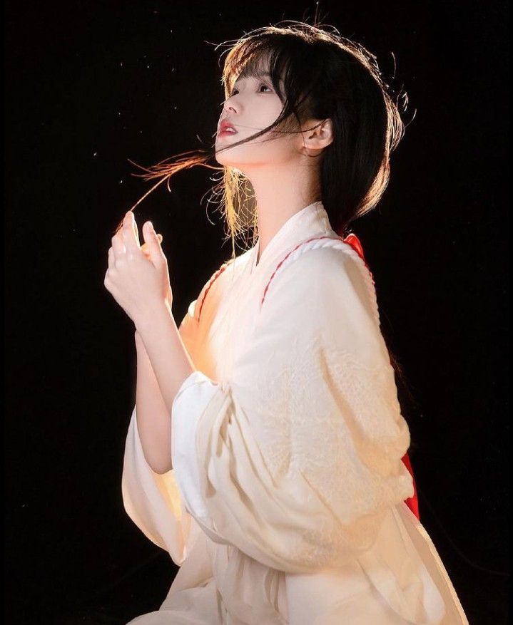 the image is a portrait of a young asian woman, possibly of chinese, japanese, or korean descent.  she's wearing a flowing, offwhite traditional garment resembling a kimono or hanfu. her dark hair is styled simply, and some strands frame her face. she's positioned in a threequarter view with her gaze slightly upward and to her left.  the background is entirely black, enhancing the contrast and drawing attention to her light clothing and the soft light illuminating her. the overall mood is serene and ethereal.  the pose suggests elegance and grace.