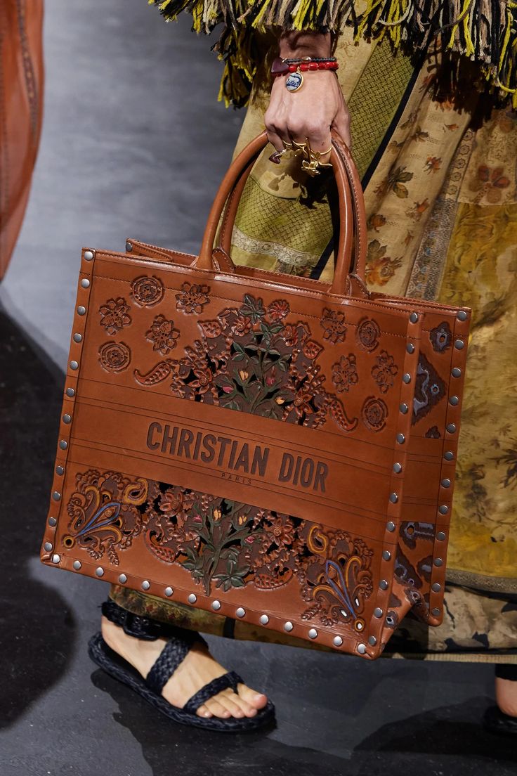 a woman wearing sandals and a long flowing patterned dress is carrying a large leather tote bag emblazoned with the christian dior logo. the bag is made of brown leather and has a cut out, floral design. it also has a metal studded design. the sandals are black and have a braided strap design.  the background is blurred. the model is walking on a runway.