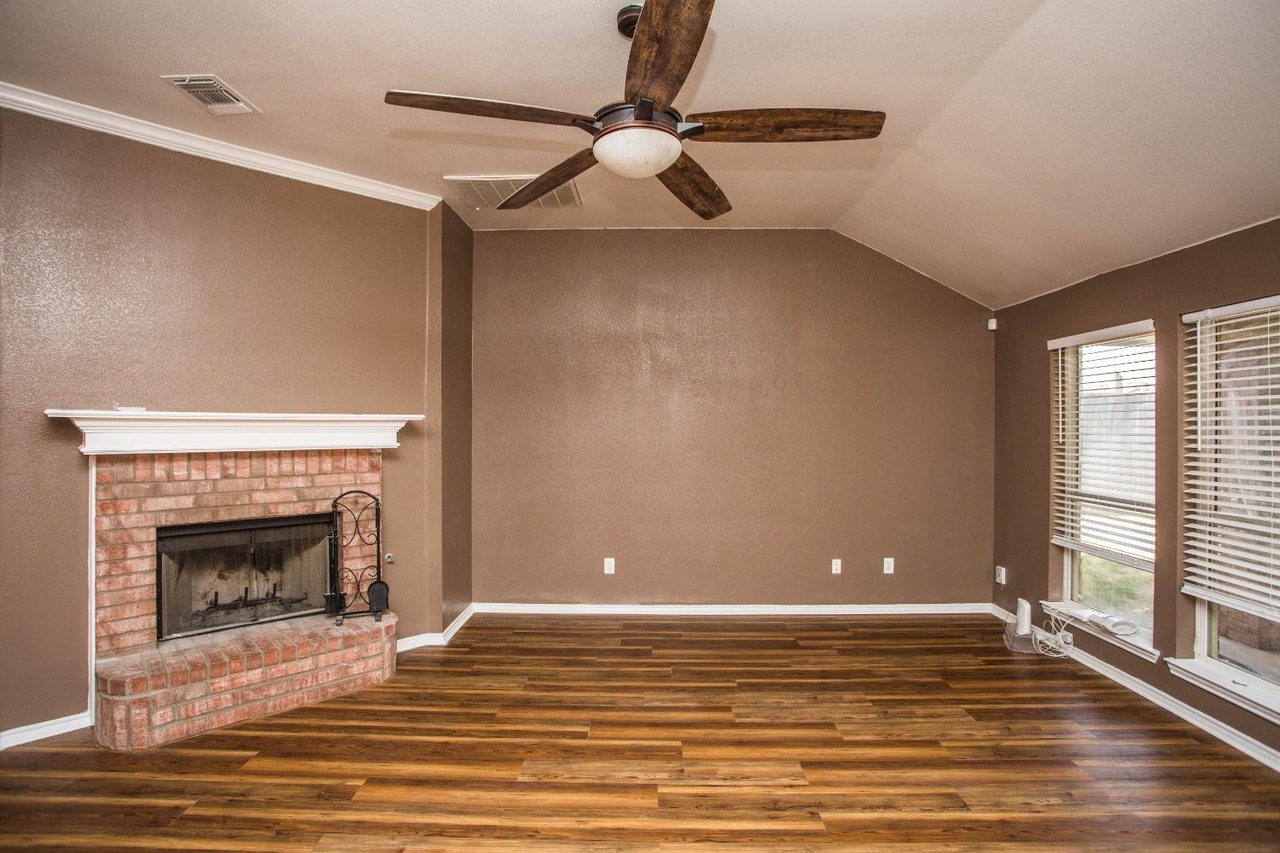 the image shows an empty living room. there is a brick fireplace on the left wall, a window with blinds on the right wall, and a ceiling fan in the middle of the ceiling. the walls are brown, and the floor is hardwood. there is white trim around the ceiling, walls, fireplace, and windows.