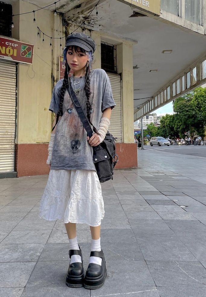 a young asian woman is standing on a city sidewalk, wearing a stylish outfit. she has on a newsboy cap, a grey wolf graphic tee, a white layered skirt, black platform mary jane shoes, and a black crossbody bag.  the overall aesthetic is a blend of grunge, indie, and alternative styles. the photo is taken outdoors in natural light.