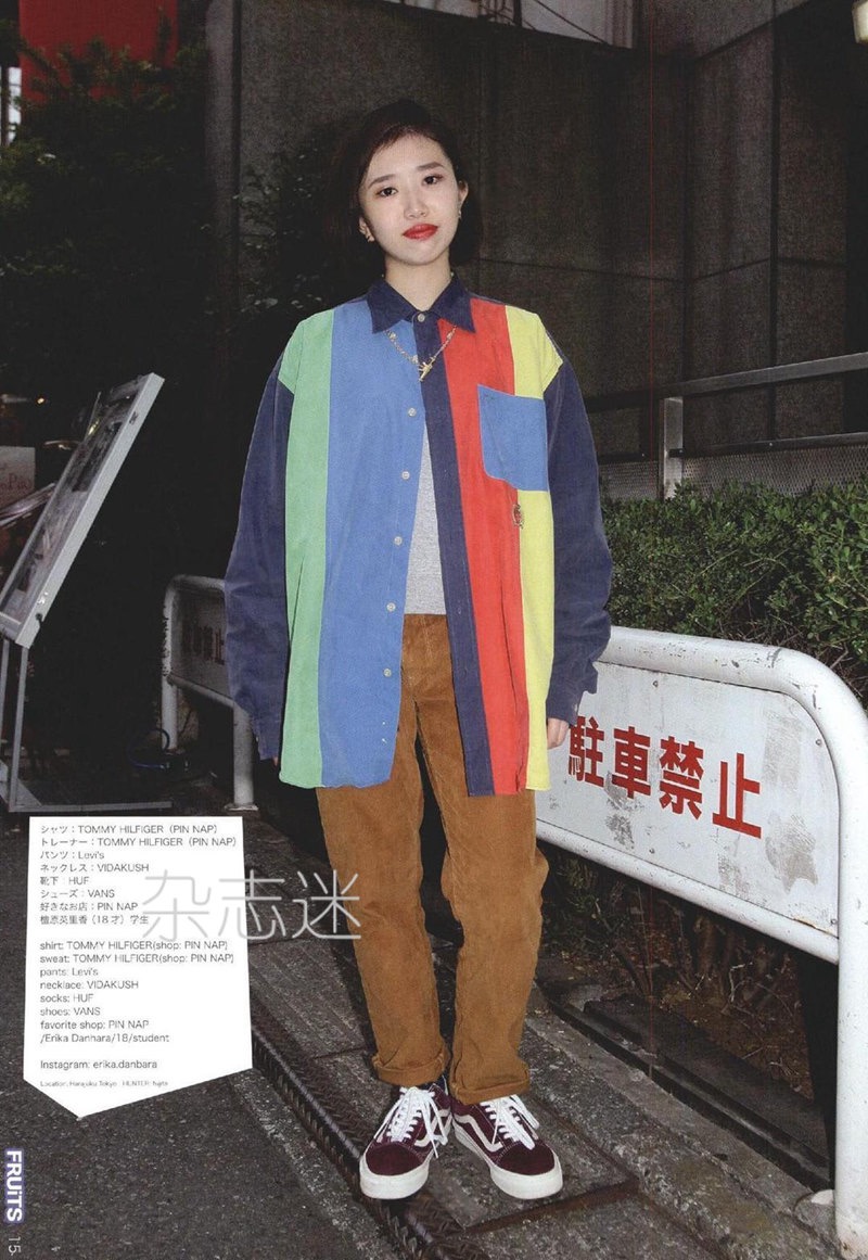 a young woman poses for a fashion photo on a city sidewalk.  she is wearing a colorful tommy hilfiger rugby shirt, brown corduroy pants, and burgundy vans sneakers. she is also wearing a gold chain. the photo has a vintage, 90s aesthetic.