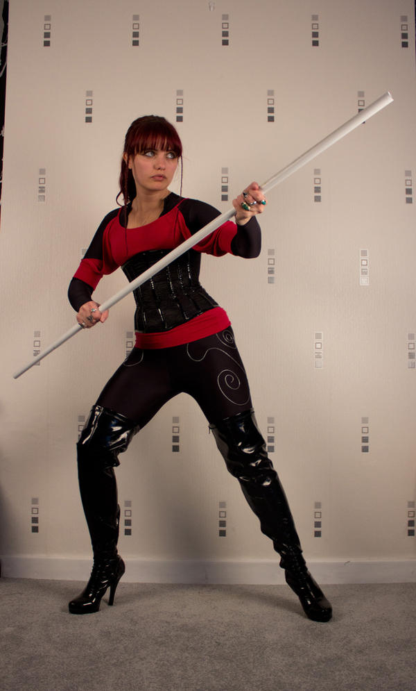 a fullbody shot of a woman dressed in a red and black martial arts costume, holding a long staff. she is wearing thighhigh black boots and has a determined expression on her face.  the background is a light grey wall with a repeating pattern.  the image suggests a dynamic action pose, possibly from a cosplay photoshoot.