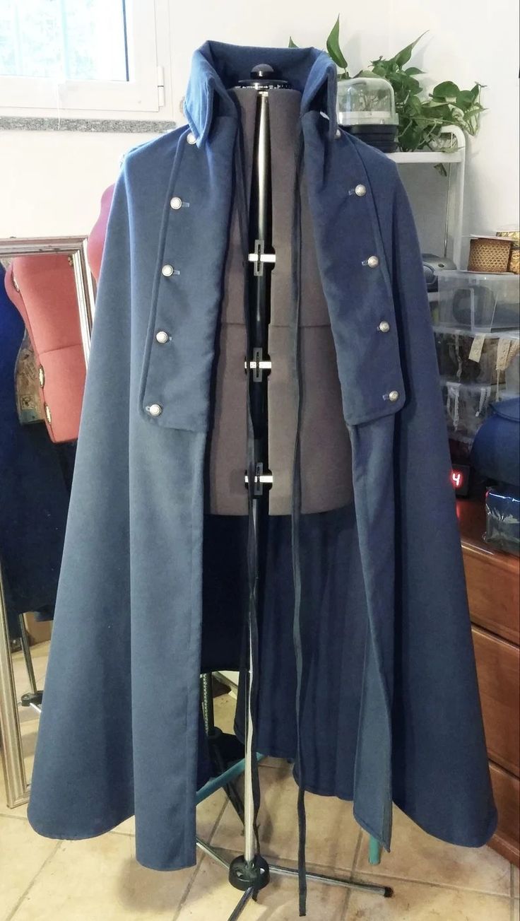 navy blue wool cape, appearing to be historically inspired (perhaps medieval or renaissance), is displayed on a mannequin.  the cape has a tailored collar, buttons down the front, and a long, flowing design. it is shown from a slightly angled front view, with a portion of the background visible including a shelf, plants, and a portion of another room.  the cape seems to be handmade and is likely a costume or piece of historicalstyle clothing.