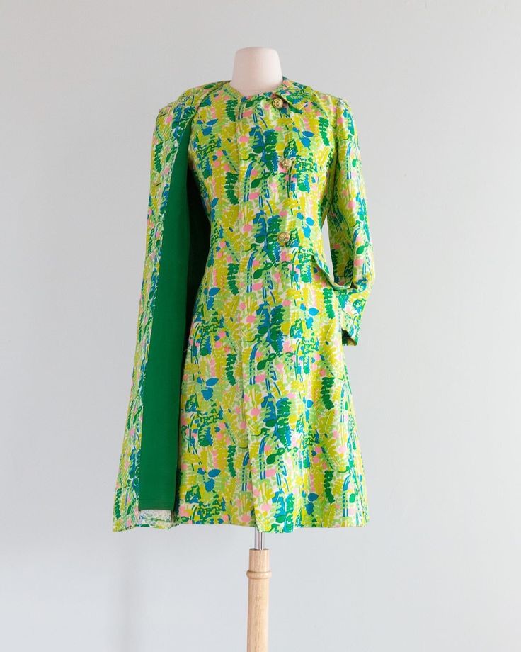 a vintage green floral shift dress with a cape is displayed on a mannequin. the dress has a colorful floral pattern in shades of green, blue, pink, and yellow. a solid green cape is attached to the dress on one side. the dress appears to be from the 1960s or 1970s era.