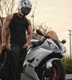 a muscular man in a black tank top and camouflage pants is standing next to a white yamaha r6 motorcycle. he is wearing a silver motorcycle helmet and is holding a black jacket in his left hand. the motorcycle is parked on the side of the road and there is a sunset in the background.