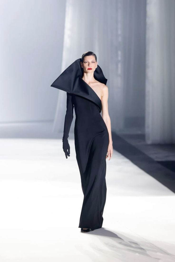 a fashion model walks down a runway in a black, oneshoulder dress with a long black sleeve and gloves. she has on natural makeup with red lipstick and her hair is styled in a chic updo.