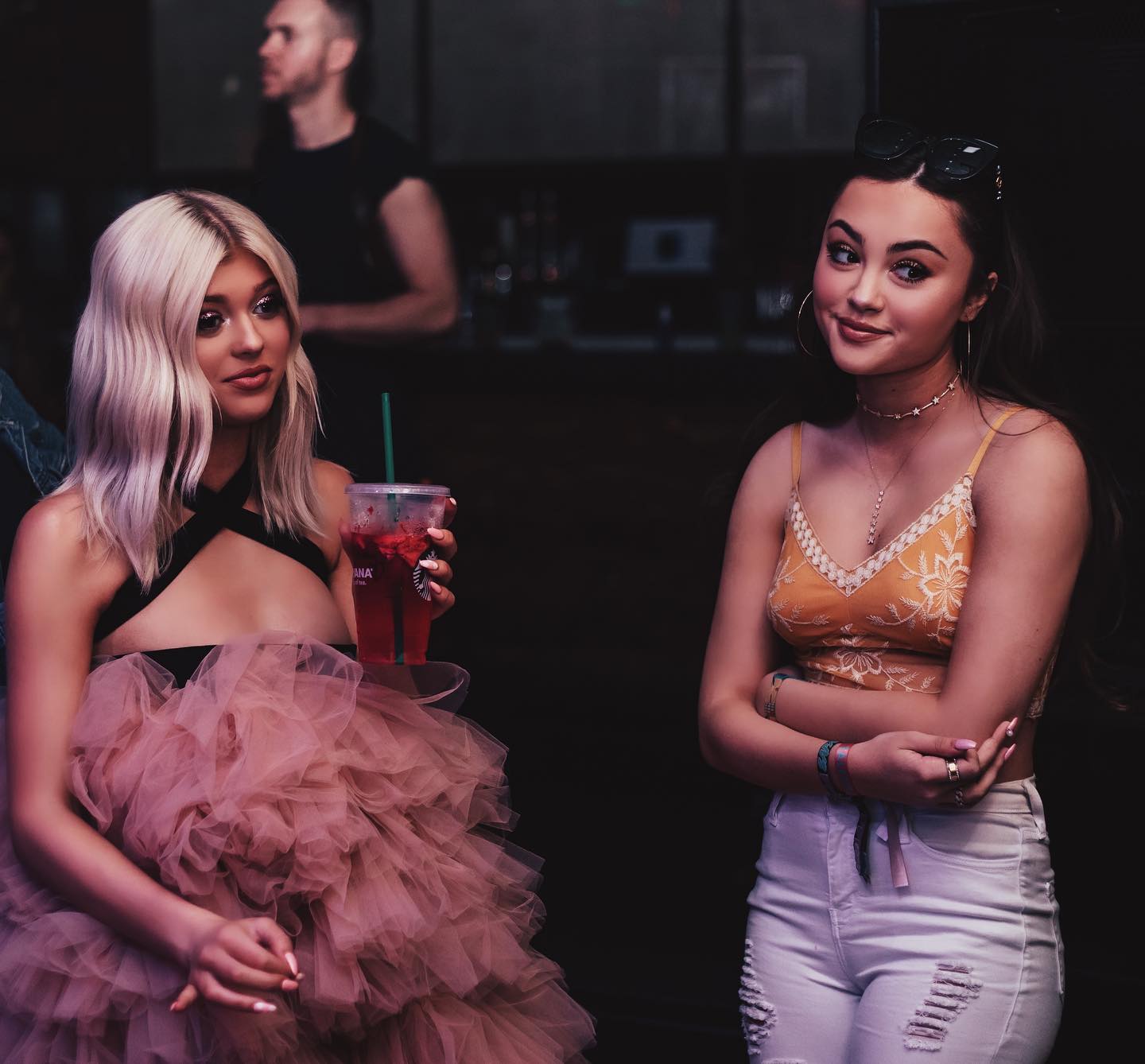 two women are standing next to each other at an event. the woman on the left has blonde hair and is wearing a black top and a large pink tulle skirt. she is holding a starbucks drink with a green straw. the woman on the right has brown hair and is wearing a yellow floral tank top and white jeans. she has sunglasses on her head and hoop earrings. there is a man standing in the background.