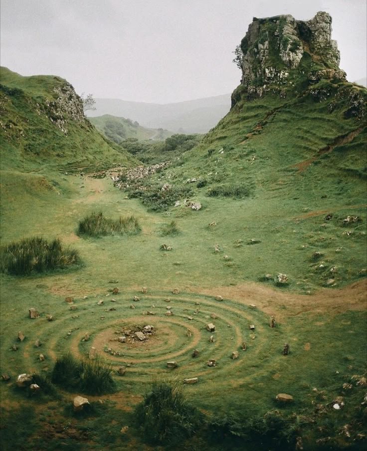 the image shows a lush, green landscape featuring rolling hills and unique rock formations. in the foreground, a spiral pattern is etched into the grass, marked by scattered stones. a valley extends into the background, with an overcast sky providing soft lighting.