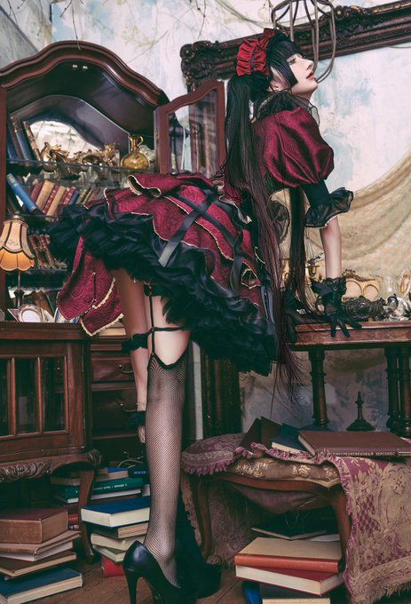 a woman dressed in a gothic lolita style outfit is posed indoors. she is wearing a short, layered red and black dress with a ruffled skirt, a red headpiece, and black pigtails. she also has on fishnet stockings, garter belts, and black high heels. she is standing near a pile of books and some dark wood furniture, including a desk and a bookcase. the setting appears to be an oldfashioned room filled with books and decorative items.
